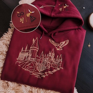 Puede incluir: Sudadera con capucha burdeos con bordados dorados de un castillo, un búho y otros símbolos. La capucha presenta gafas bordadas, una varita, un rayo y las palabras "9 3/4" y "HP".