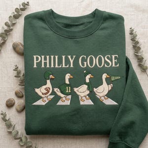 Może przedstawiać: Ciemnozielona bluza z okrągłym dekoltem i białym napisem PHILLY GOOSE. Poniżej wyhaftowano cztery białe gęsi idące po białych pasach. Gęsi mają na sobie kask do futbolu amerykańskiego, zieloną koszulkę z numerem 11, szalik oraz trzymają małą flagę.
