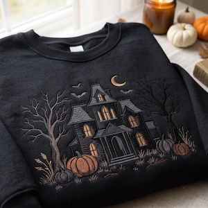 Pode incluir: Uma camisola preta de gola redonda com bordado detalhado de uma casa assombrada, árvores despidas e abóboras. O design inclui uma lua crescente e morcegos a voar em tons de laranja, cinzento e preto, criando uma estética sazonal.