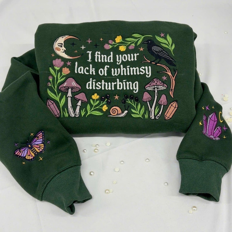May include: A dark green sweatshirt featuring an embroidered design with the text "I find your lack of whimsy disturbing." The graphic includes a crescent moon, a black crow, mushrooms, a snail, flowers, and crystals. A butterfly is embroidered on the left sleeve and crystals on the right sleeve.