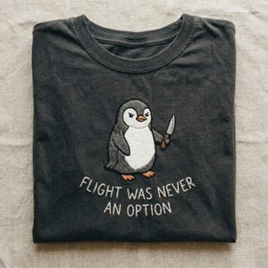 Peut inclure: Un t-shirt &agrave; col rond gris anthracite avec une broderie repr&eacute;sentant un pingouin tenant un petit couteau. Sous le pingouin, le texte FLIGHT WAS NEVER AN OPTION est brod&eacute; en fil blanc. Le motif est centr&eacute; sur la poitrine du v&ecirc;tement.
