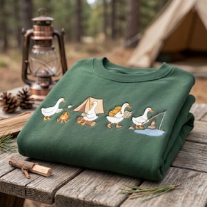 Può includere: Una felpa verde foresta piegata, con un ricamo di oche in campeggio. Le oche sono raffigurate mentre arrostiscono marshmallow, montano una tenda, trasportano uno zaino e pescano. La felpa è su una superficie di legno.