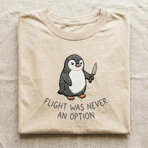 Peut inclure: Un t-shirt beige &agrave; col rond avec une broderie repr&eacute;sentant un manchot tenant un petit couteau. Sous le manchot, le texte FLIGHT WAS NEVER AN OPTION est brod&eacute; en fil gris fonc&eacute;. Le t-shirt est compos&eacute; d'un tissu beige clair uni.