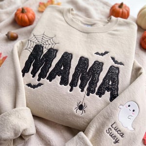 Könnte beinhalten: Ein beigefarbenes Sweatshirt mit Rundhalsausschnitt und dem Wort MAMA in schwarzen, glitzernden, tropfenden Buchstaben. Das Design umfasst ein Spinnennetz, eine Spinne und schwarze Fledermäuse. Der Ärmel zeigt ein gesticktes weißes Gespenst mit den Namen Luca und Susy.
