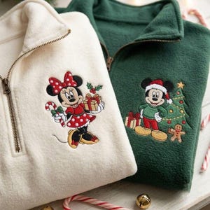 May include: A cream-colored fleece quarter-zip pullover featuring an embroidered Minnie Mouse holding a candy cane and gift, alongside a dark green fleece quarter-zip pullover featuring an embroidered Mickey Mouse in a Santa hat next to a Christmas tree.