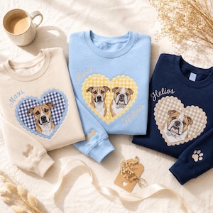 May include: Three crewneck sweatshirts in cream, light blue, and navy blue. Each features an embroidered heart with a gingham pattern containing a dog portrait. The cream sweatshirt shows a dog named Maxi, the blue shows two dogs, and the navy shows Helios.