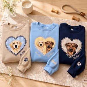 May include: Three crewneck sweatshirts in beige, light blue, and navy blue. Each features a heart-shaped patch with dog illustrations and names Maxi and Helios. The sleeves include embroidered paw prints or bone shapes on the cuffs.