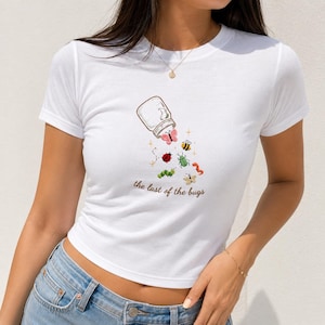 May include: A white short-sleeved t-shirt featuring a graphic of a glass jar spilling out various colourful insects and a butterfly. Below the illustration, the text reads "the last of the bugs" in a brown, cursive, lowercase font.