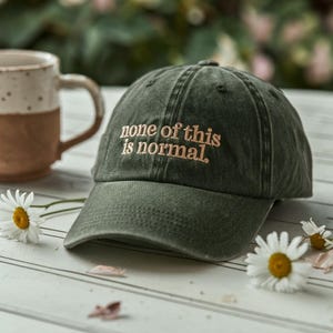 May include: A dark green baseball cap with the embroidered text "none of this is normal." The hat is sitting on a white surface with a daisy and a mug in the background. The cap has a curved brim.