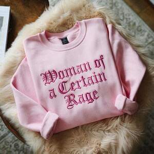 May include: A light pink sweatshirt with the embroidered text "Woman of a Certain Rage" in a gothic font. The sweatshirt has a crew neck and long sleeves with rolled cuffs. The garment is displayed on a fluffy, light-colored surface.