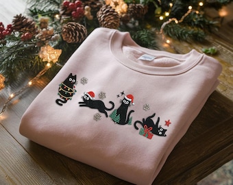 May include: A pale pink sweatshirt with a Christmas cat design. The black cats are adorned with festive elements like Santa hats, Christmas lights, and snowflakes. A seasonal sweatshirt for the holidays.