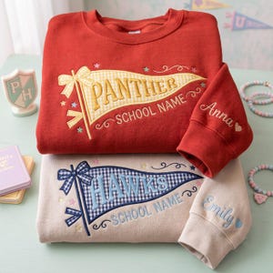 Puede incluir: Una sudadera roja con un banderín de cuadros amarillos bordado que dice PANTHER SCHOOL NAME y una sudadera beige con un banderín de cuadros azules que dice HAWKS SCHOOL NAME. Las mangas tienen bordados los nombres Anna y Emily.