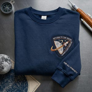 Op de afbeelding: Marineblauw sweatshirt met een ronde hals en geborduurde patch. De patch luidt "Return to the Moon Lunar Flyby Mission" met een raket- en maanontwerp. De mouw luidt "For All Humanity".