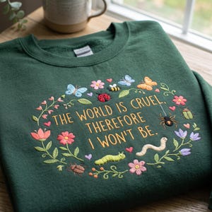 May include: A dark green crewneck sweatshirt featuring embroidered text that reads, THE WORLD IS CRUEL, THEREFORE I WON'T BE. The text is surrounded by a colorful embroidered wreath of various flowers, small hearts, butterflies, a bee, a ladybug, a spider, and caterpillars.
