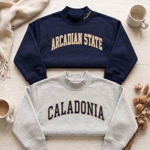 Könnte beinhalten: Ein marineblaues Sweatshirt mit Stehkragen und dem Text ARCADIAN STATE in Gold sowie ein hellgraues Sweatshirt mit Stehkragen und dem Text CALADONIA in Marineblau. Beide Kleidungsstücke haben lange Ärmel und gerippte Kragen, mit Arcadians am Hals.