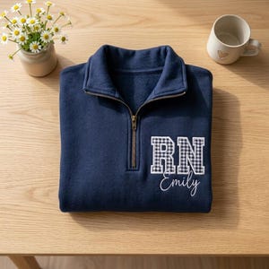 May include: A navy blue quarter-zip sweatshirt with a gold zip. The sweatshirt displays the letters "RN" in a black and white checked pattern, with the name "Emily" embroidered below in white script. A mug and flowers are in the background.