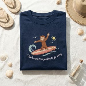 May include: Navy blue t-shirt featuring a cartoon monkey surfing on a surfboard, with the text "I don't want this feeling to go away." The design includes a sun, waves, and seagulls. The shirt is surrounded by beach-themed accessories.
