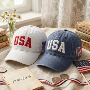May include: Two baseball caps, one white with red embroidered USA text and one blue with white embroidered USA text. Both caps feature a small American flag patch on the side with the text 1776-2026. The caps have a structured crown and a curved brim.