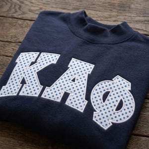 Op de afbeelding: Een marineblauw sweatshirt met ronde hals en grote, witte Griekse letters in blokstijl met een blauw stippenpatroon. De letters K, A en Phi zijn op de borst aangebracht met een witte gestikte rand, wat zorgt voor een textuur.