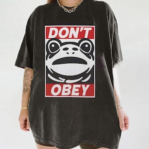 May include: Dark gray t-shirt featuring a stylized frog design with the words "DON'T OBEY" in a red and white rectangular frame. The shirt has short sleeves and a relaxed fit, suitable for casual wear.
