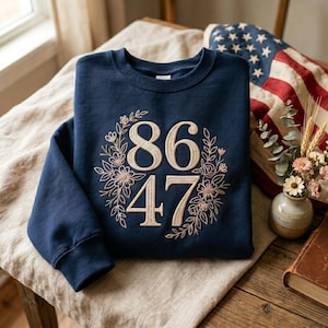 May include: A navy blue sweatshirt with the numbers "86" and "47" embroidered in a light beige color, surrounded by a floral wreath. The sweatshirt is folded on a beige linen surface, with an American flag in the background.