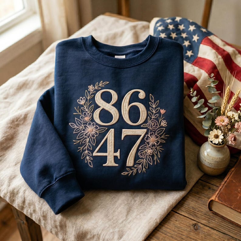 May include: A navy blue sweatshirt with the numbers "86" and "47" embroidered in a light beige color, surrounded by a floral wreath. The sweatshirt is folded on a beige linen surface, with an American flag in the background.