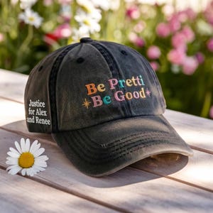 May include: A faded black baseball cap embroidered with the words "Be Pretti Be Good" in various colours. The cap also has the text "Justice for Alex and Renee" on the side. A daisy lies on a wooden surface.