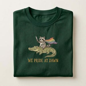 May include: Dark green t-shirt with a cartoon raccoon riding an alligator, holding a sword and wearing a rainbow cape. The text "WE PRIDE AT DAWN" is printed in gold. The shirt is folded. The design is whimsical.