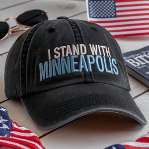 以下が含まれることがあります： ライトピンクとブルーで「I STAND WITH MINNEAPOLIS」と刺繍された黒い野球帽。帽子は使い古された生地で作られており、つばがカーブしています。サングラスとアメリカ国旗が背景にあります。