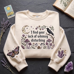 May include: A cream-colored crewneck sweatshirt featuring a central graphic with the text "I find your lack of whimsy disturbing," surrounded by a crescent moon, a crow, mushrooms, flowers, and a snail. The sleeves feature embroidered moth and crystal designs.