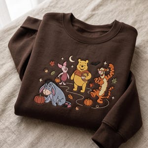 Könnte beinhalten: Ein dunkelbraunes Sweatshirt mit Rundhalsausschnitt und einer Stickerei von Winnie Puuh, Ferkel, Tigger und I-Aah. Das Motiv zeigt herbstliche Elemente wie Kürbisse, Ahornblätter, Eicheln, eine Mondsichel, Sterne und eine Fledermaus.