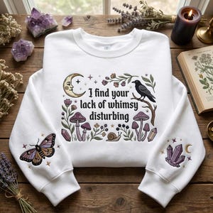 May include: A white crewneck sweatshirt featuring a central graphic with the text 'I find your lack of whimsy disturbing' surrounded by a moon, a raven, mushrooms, and a snail. The sleeves feature embroidered moth and crystal designs.