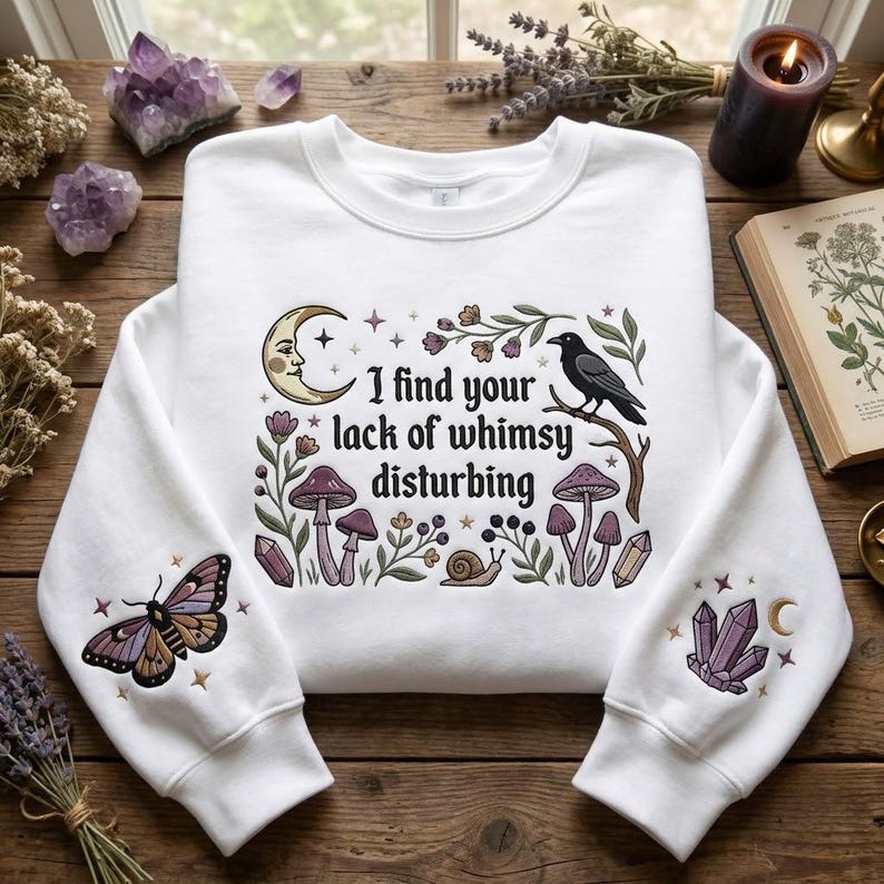 May include: A white crewneck sweatshirt featuring a central graphic with the text 'I find your lack of whimsy disturbing' surrounded by a moon, a raven, mushrooms, and a snail. The sleeves feature embroidered moth and crystal designs.