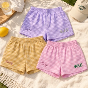 May include: Three pairs of elastic-waist shorts in gingham patterns. One pair is purple with the name Sophie and Greek letters KΔΣ embroidered. One pair is yellow with the name Avery. One pair is pink with the name Maya and Greek letters ΦΛΕ.