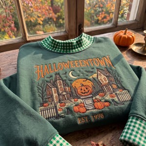 May include: A dark green sweatshirt with a green and white gingham collar and cuffs. The front features an embroidered scene of two houses, a crescent moon, and pumpkins with the text HALLOWEENTOWN and EST. 1998 in orange thread.