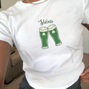 May include: White t-shirt with a green graphic of two beer glasses with shamrocks and the word "Sláinte" in a script font. The beer glasses have foamy tops. The shirt is short-sleeved.