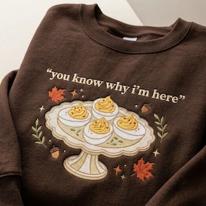 May include: A dark brown crewneck sweatshirt featuring an embroidered design of four deviled eggs on a pedestal platter. The text "you know why i'm here" is embroidered above the platter, surrounded by small autumn leaves, acorns, and stars.