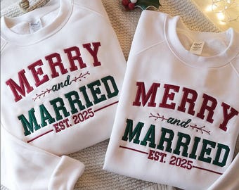 Può includere: Due felpe bianche con le parole "MERRY and MARRIED EST. 2025" ricamate in rosso e verde. Le felpe sono su una superficie neutra, con decorazioni natalizie sullo sfondo.