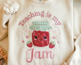 May include: Cream-colored sweatshirt with the embroidered phrase "Teaching is my Jam." The design features a smiling jar of red jam with a green gingham lid, surrounded by stars and strawberries. The text and images are in pink.