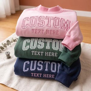 Puede incluir: Una pila de tres sudaderas de manga larga en rosa, verde oscuro y azul marino. Cada sudadera presenta un cuello alto, puños acanalados y el texto bordado CUSTOM con un patrón de cuadros, seguido del texto TEXT HERE debajo.