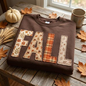 May include: A dark brown crewneck sweatshirt featuring the word FALL appliqued across the front. Each letter is crafted from fabric with distinct patterns, including floral prints, acorn motifs, and an orange plaid design with visible stitching.