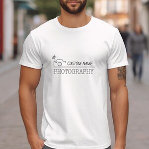 Può includere: Maglietta bianca con un'icona di una fotocamera nera e il testo "CUSTOM NAME PHOTOGRAPHY" stampato sul davanti.