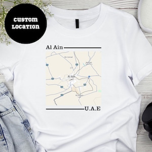 Puede incluir: Camiseta blanca con un mapa en blanco y negro de Al Ain, Emiratos Árabes Unidos, impreso en la parte delantera. El texto "Al Ain" y "E.A.U." está impreso debajo del mapa.