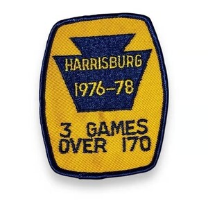May include: A yellow embroidered patch with a dark blue border and a central dark blue keystone shape. The text reads HARRISBURG 1976-78 in yellow and 3 GAMES OVER 170 in dark blue. The patch has a rounded rectangular shape with stitched edges.