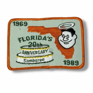 May include: A rectangular embroidered patch with an orange border on a light green background. It features an orange map of Florida, a white two-tier cake, and a cartoon face with a halo. Text includes 1969, 1989, and Florida's 20th Anniversary Samboree.