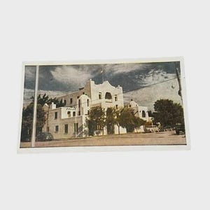May include: A vintage postcard featuring a multi-story white building with a flat roof and arched architectural details. The structure is surrounded by green trees and a paved street with parked cars under a cloudy sky in a muted color palette.