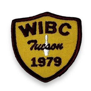 May include: A shield-shaped embroidered patch with a gold background and a dark brown border. The text reads WIBC, Tucson, and 1979 in dark brown lettering. A white bowling pin graphic is centered between the text lines on the patch surface.