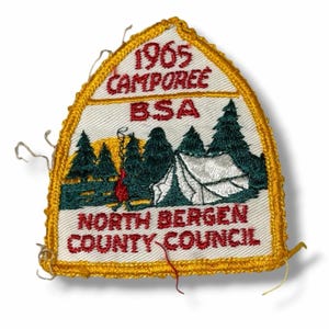 May include: A shield-shaped embroidered patch with a yellow border. It features a white background with red text reading 1965 CAMPOREE BSA and NORTH BERGEN COUNTY COUNCIL. The center depicts a white tent, green pine trees, and a red campfire.