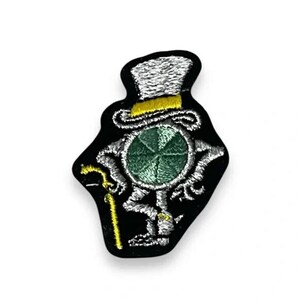May include: An embroidered patch featuring a circular green center with white stitched limbs, wearing a white top hat with a yellow band. The figure has a white stitched cane in one hand and is set against a solid black background shape.