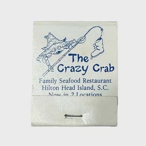May include: A white matchbook cover featuring a blue line drawing of a crab wearing a hat and fishing for a boot. The text reads The Crazy Crab, Family Seafood Restaurant, Hilton Head Island, S.C., Now in 2 Locations, with a metal staple visible.